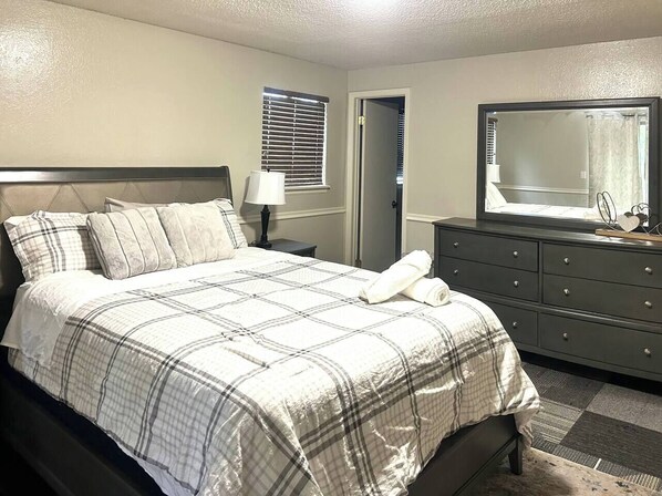 Une chambre à coucher comprenant un lit, une commode, un miroir, une table de chevet et une fenêtre avec des stores.