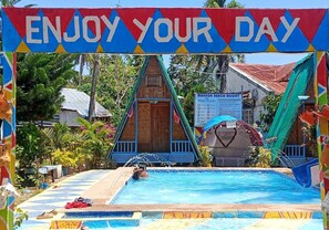 A pool area with a sign that reads 'ENJOY YOUR DAY,' a wooden structure, and a banner for 'MANNOR BEACH RESORT.'