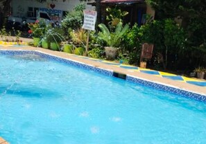 A swimming pool with a tiled edge, surrounded by greenery and a building with a sign.