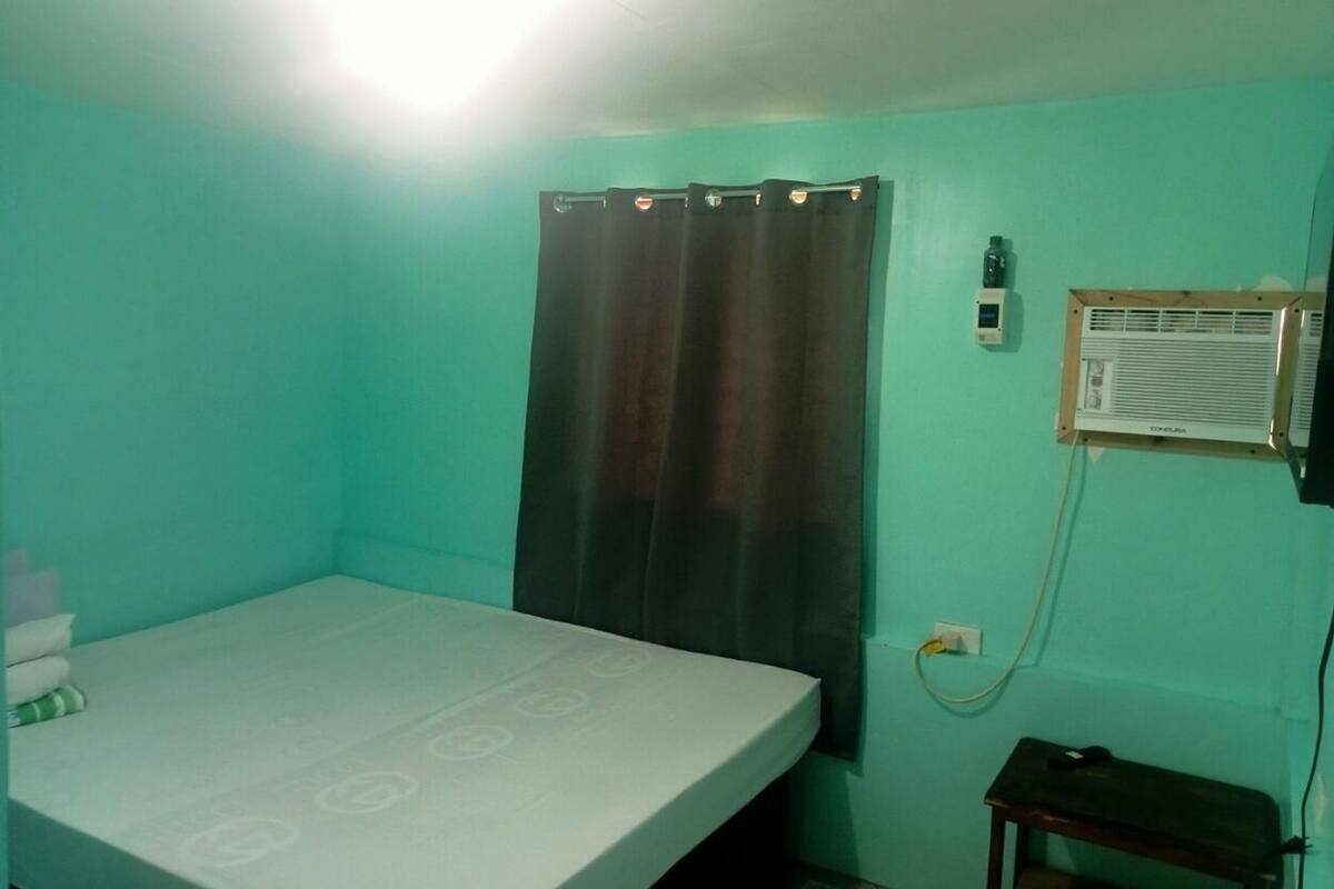 A room with teal walls, a bed with a patterned bedspread, a curtain, a wall-mounted air conditioner, and a small table.