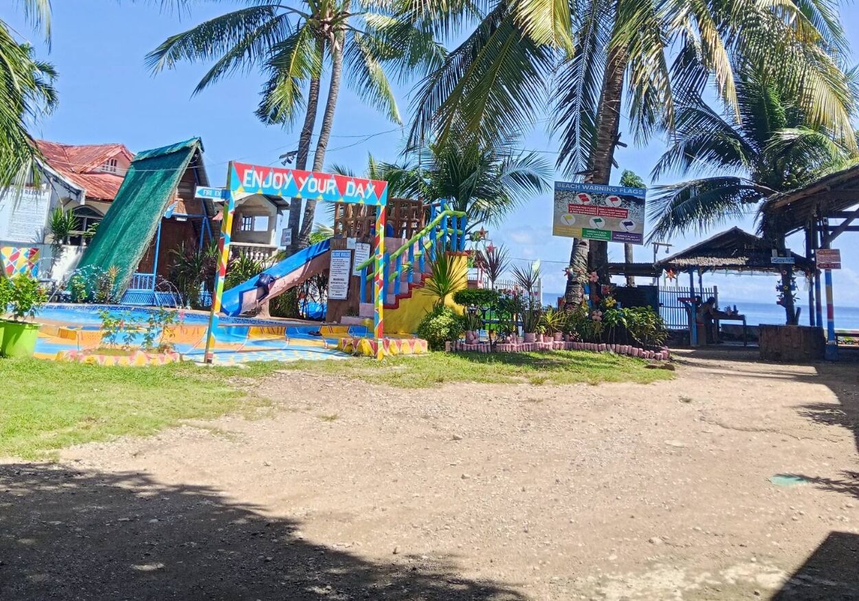 A colorful playground with a sign that reads 'ENJOY YOUR DAY,' surrounded by palm trees and a beach view.