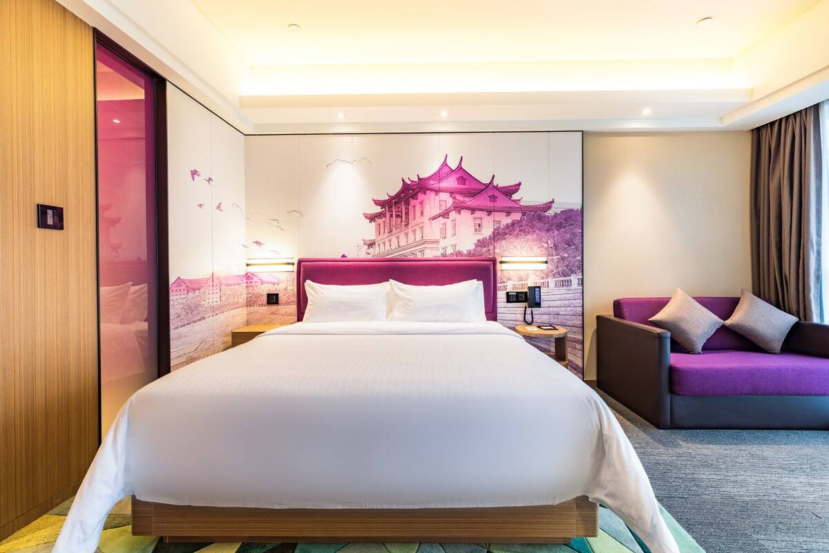 Ein Hotelzimmer mit einem großen Bett, einem violetten Sofa, einem Wandbild und einer Holztür.