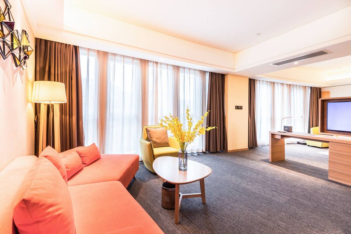 Ein Hotelzimmer mit einer Couch, einem kleinen Tisch mit Blumenvase, einem Schreibtisch mit Computer und einem Fernseher.
