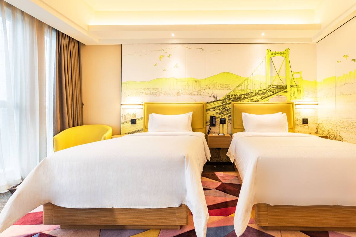Ein Hotelzimmer mit zwei Betten, einem gelben Sessel, einem großen Wandbild und einem gemusterten Teppich.