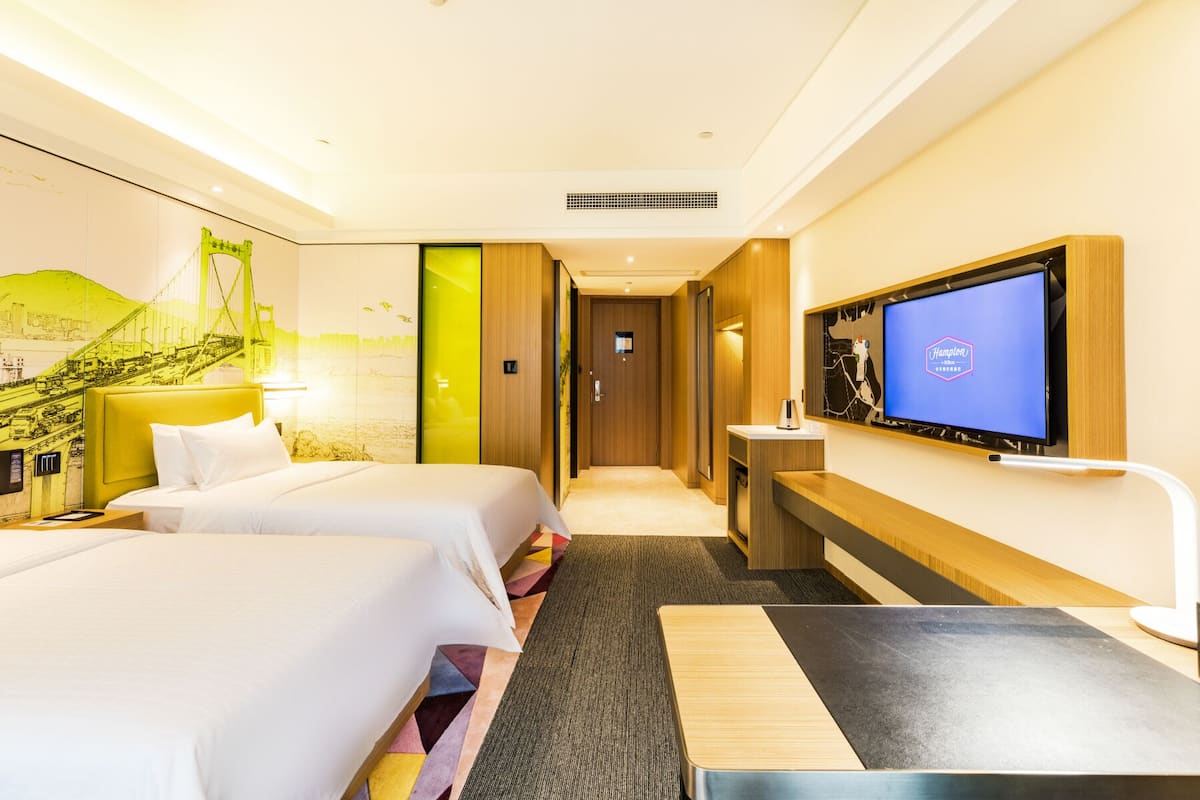 Ein Hotelzimmer mit zwei Betten, einem Schreibtisch, einem Fernseher und einem Wandgemälde einer Brücke.
