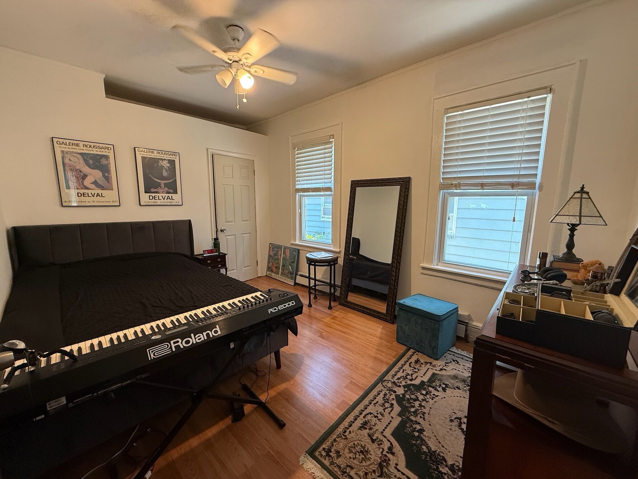 A bedroom with a bed, a standing keyboard, a ceiling fan, a mirror, and a window with blinds.