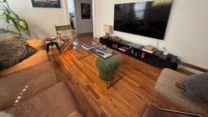 Een woonkamer met een houten vloer, een grote flatscreen televisie, een groene poef, een leren bank en een regisseurstoel.