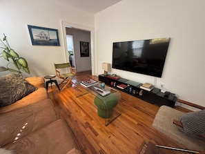 Ein Wohnzimmer mit Holzboden, einem großen Flachbildfernseher, einem grünen Hocker, einer Ledersofa und einem Regiestuhl.