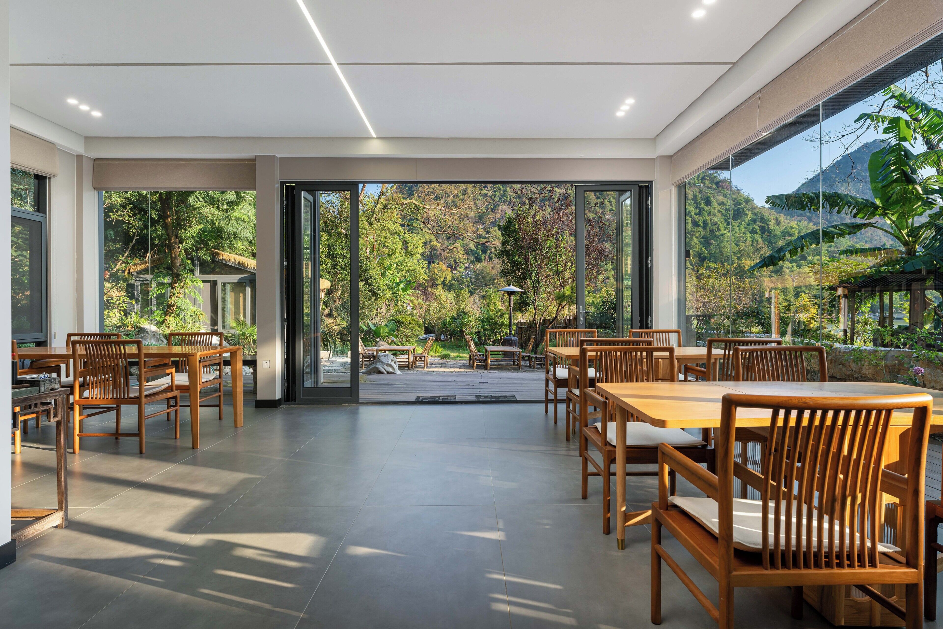 A spacious indoor dining area with wooden tables and chairs, large windows offering a view of a lush garden and mountains.