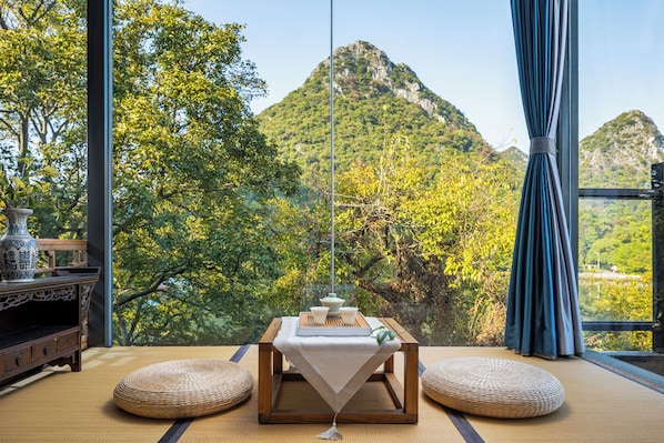 A room with tatami flooring, a low wooden table with a white cloth, and a view of a mountainous landscape.