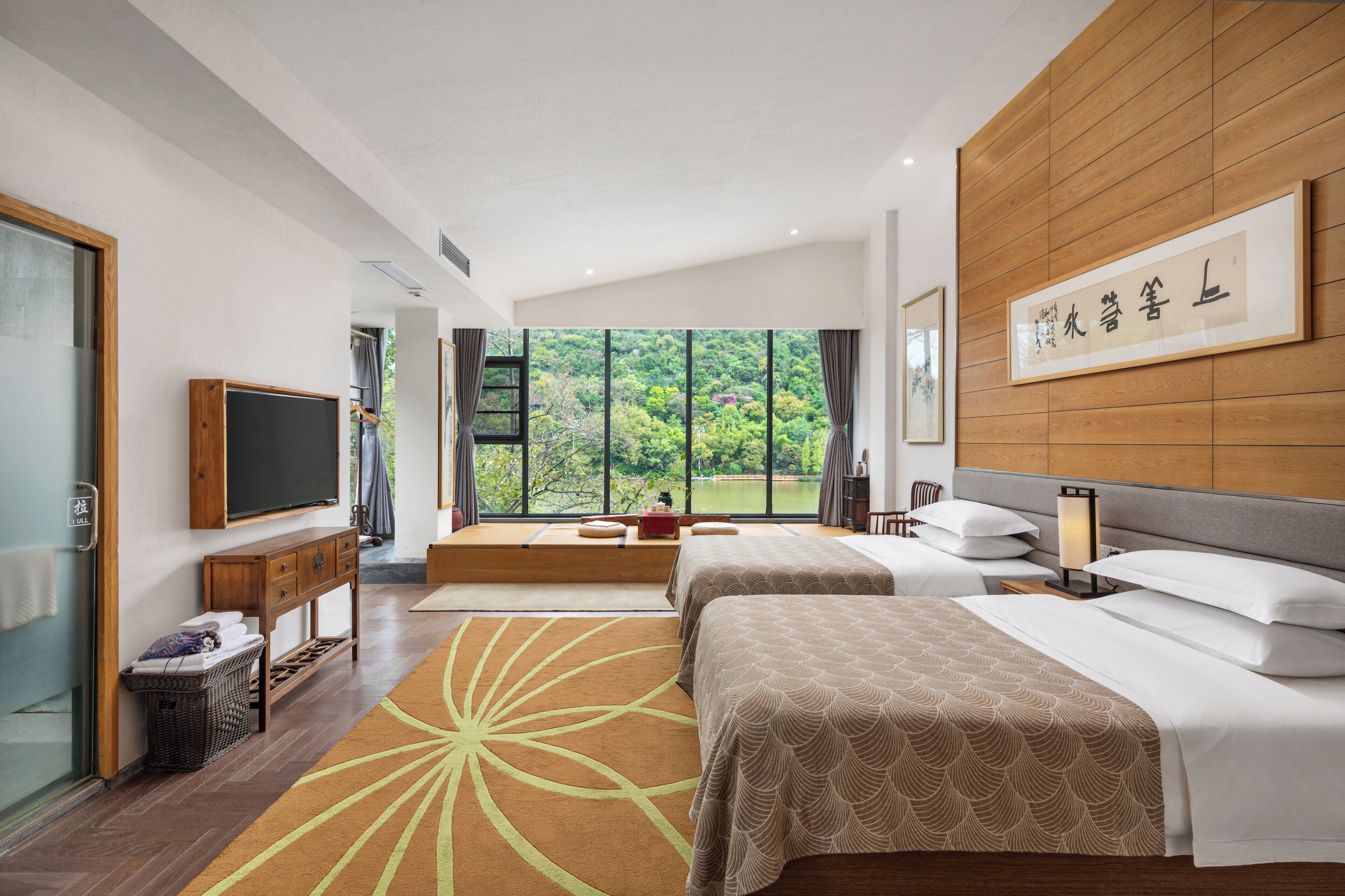 A modern hotel room with a large window, a flat-screen TV, a bed with a patterned bedspread, a bedside table with a lamp, and a wooden headboard.