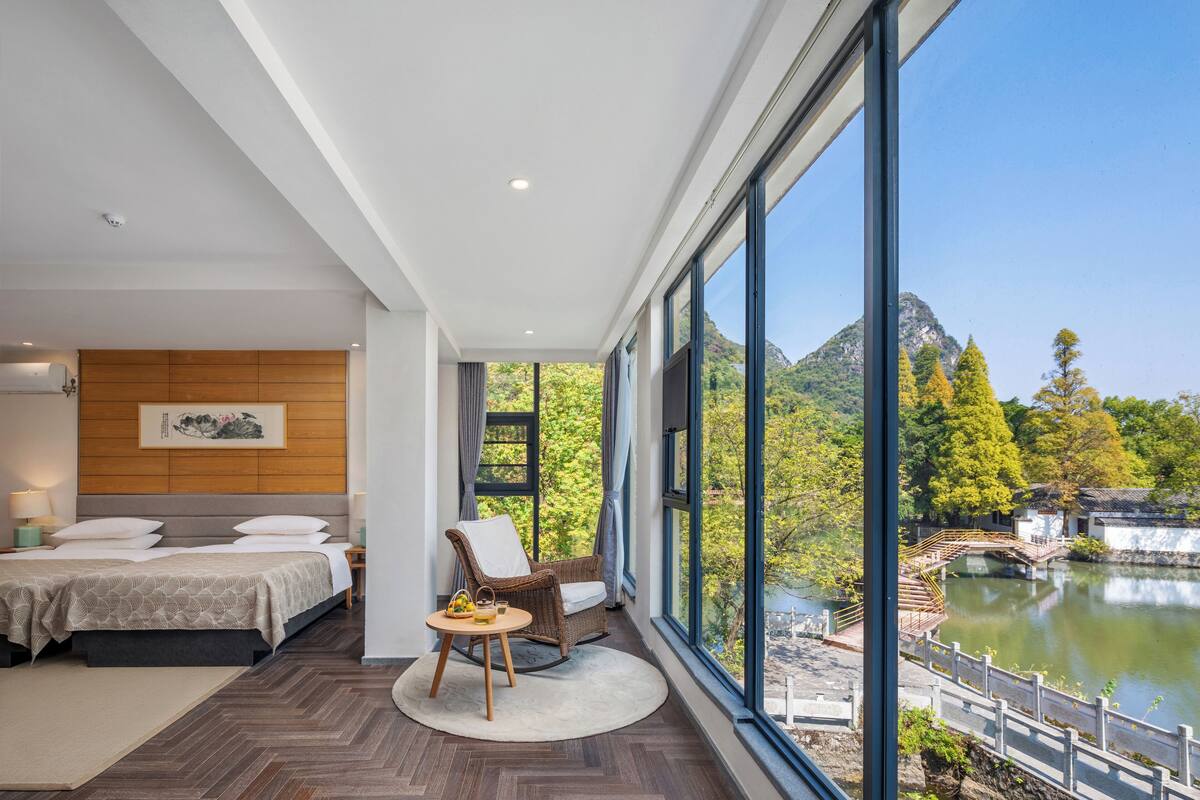 A modern bedroom with a large window, a bed, a chair, and a view of a lake and mountains.