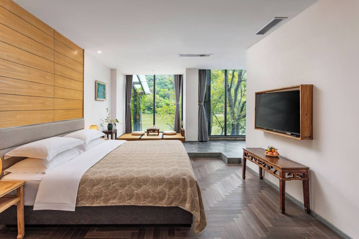 A modern hotel room with a large bed, a TV, a wooden bench, and a view of trees through the windows.
