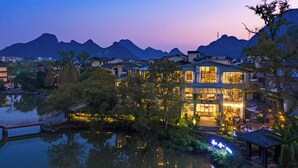 A modern hotel with a large glass facade, a courtyard, and a view of mountains. - Guilin Heshe Lakeside Resort (Guilin)