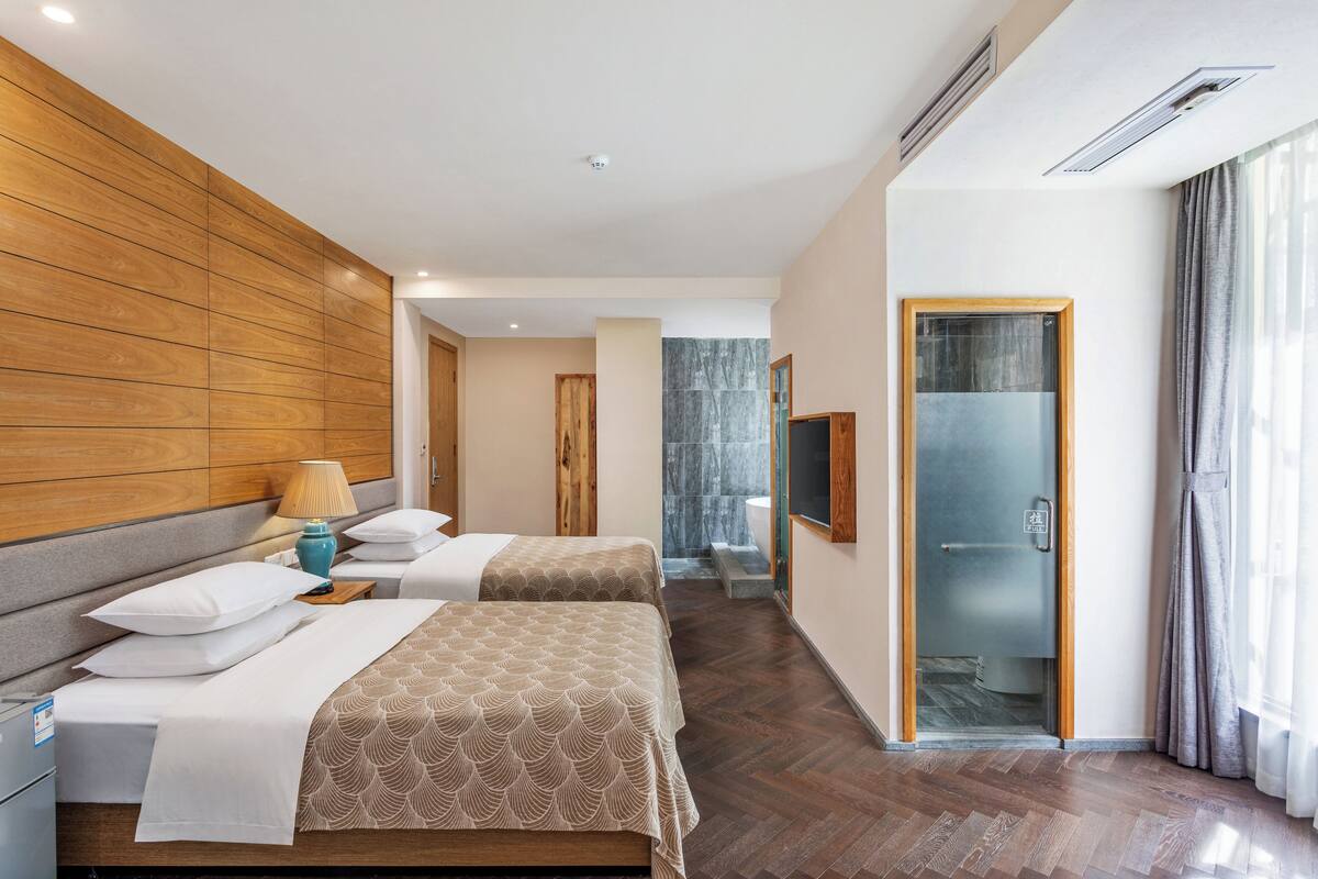 A hotel room with two beds, a wooden headboard, a bathroom with a glass shower, and a view of the outside.