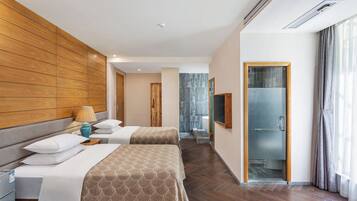 Quarto de hotel com duas camas, cabeceira de madeira, banheiro com chuveiro de vidro e vista para o exterior.