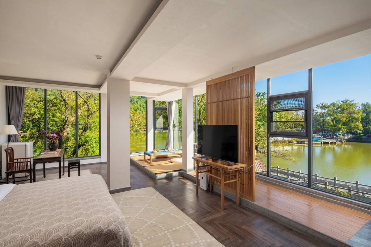 A hotel room with a large bed, a TV, a desk with a chair, and a view of a lake.