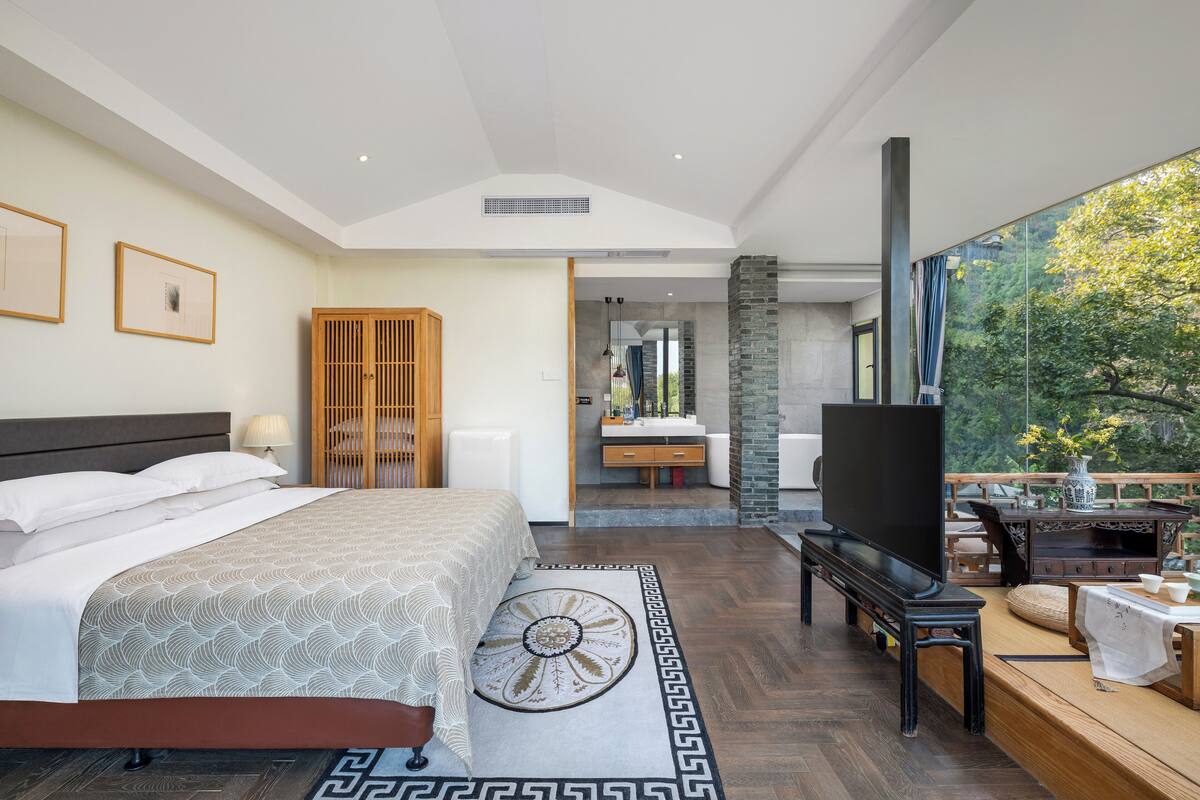 A modern bedroom with a bed, a TV, a wooden bench, and a view of a bathroom and outdoor area.