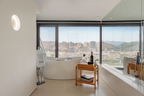 A modern hotel room with a large window offering a city view, a small wooden cart with books, and a white reception desk.