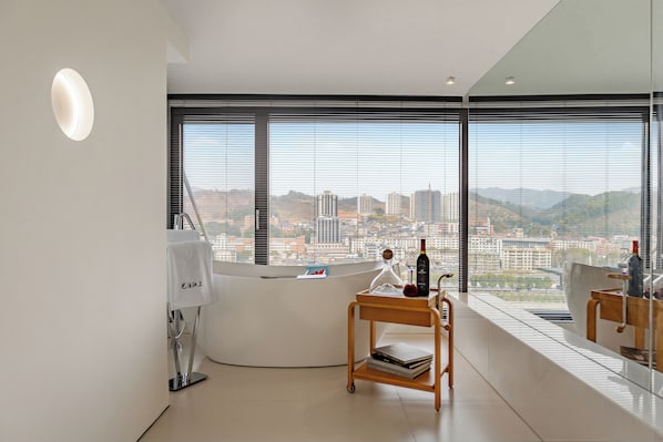 A modern hotel room with a large window offering a city view, a small wooden cart with books, and a white reception desk.
