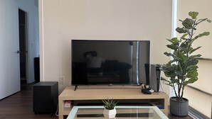 A living room with a flat-screen TV on a wooden stand, a glass coffee table, a potted plant, and a door leading to another room.