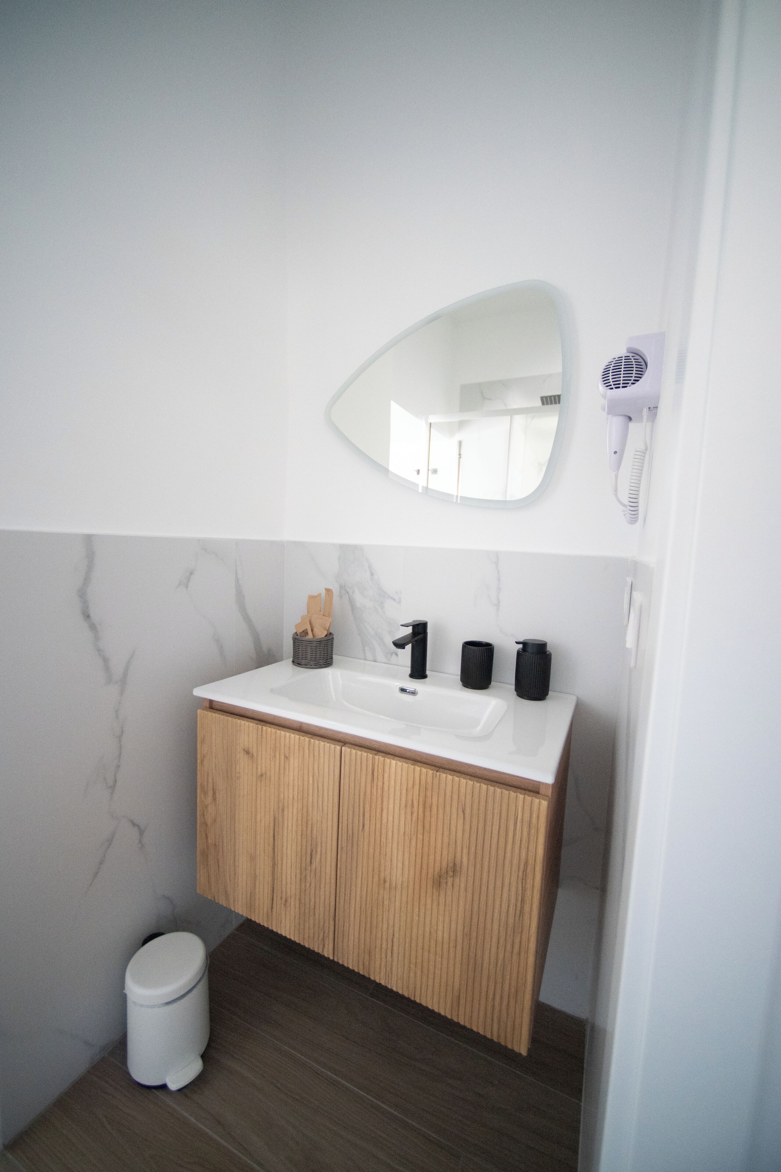 Een moderne badkamer met een marmeren wand, een houten wastafelmeubel, een witte wasbak en een aan de muur bevestigde haardroger.