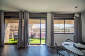 Ein Zimmer mit einer Glasschiebetür, die auf einen Balkon mit Blick auf ein Gebäude und einen umzäunten Garten führt. Auf dem Tisch stehen Stühle und eine Fernbedienung.