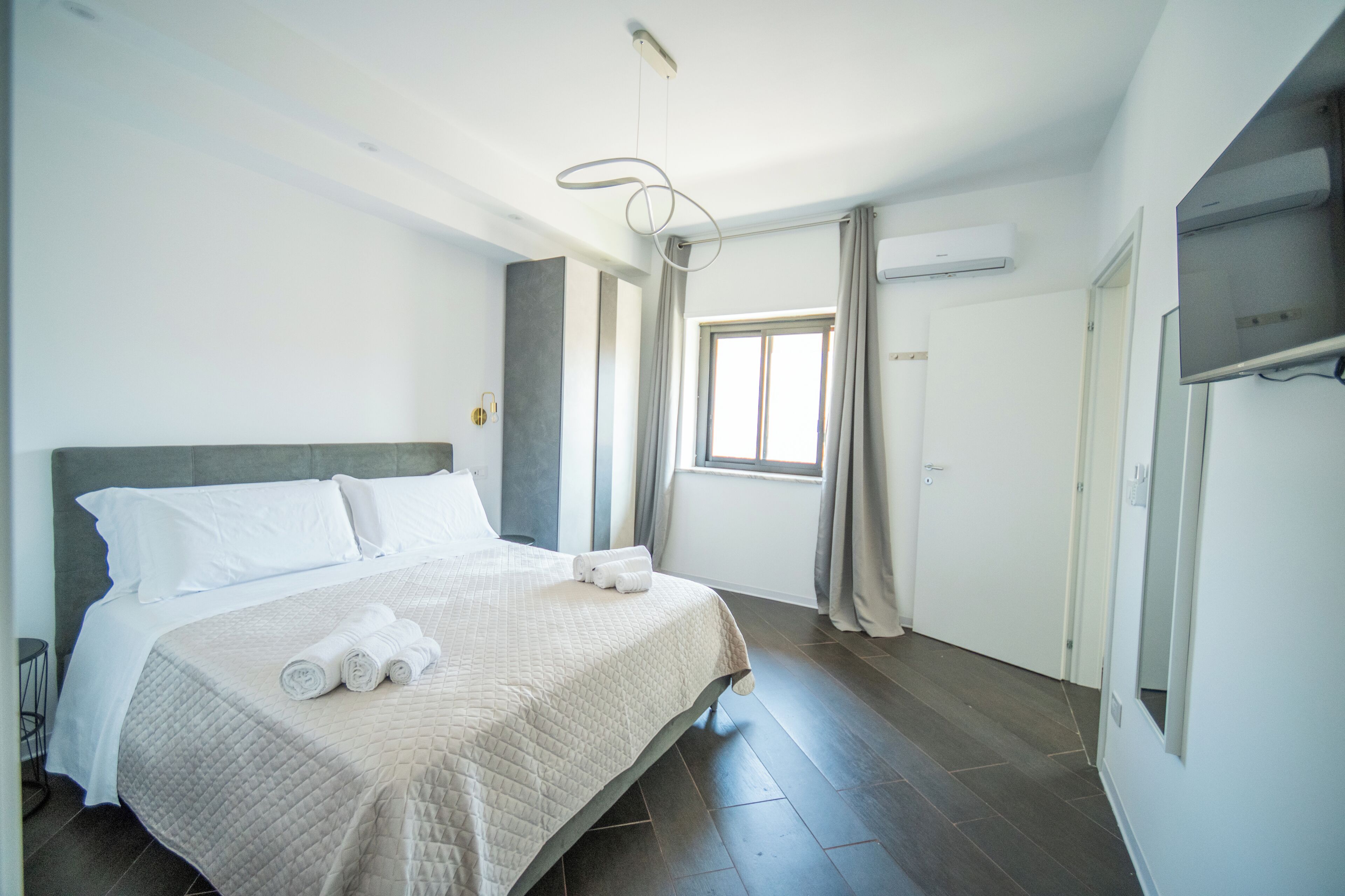 Ein Hotelzimmer mit einem großen Bett, einem Nachttisch, einem Fenster mit Vorhängen und einer fest installierten Klimaanlage.