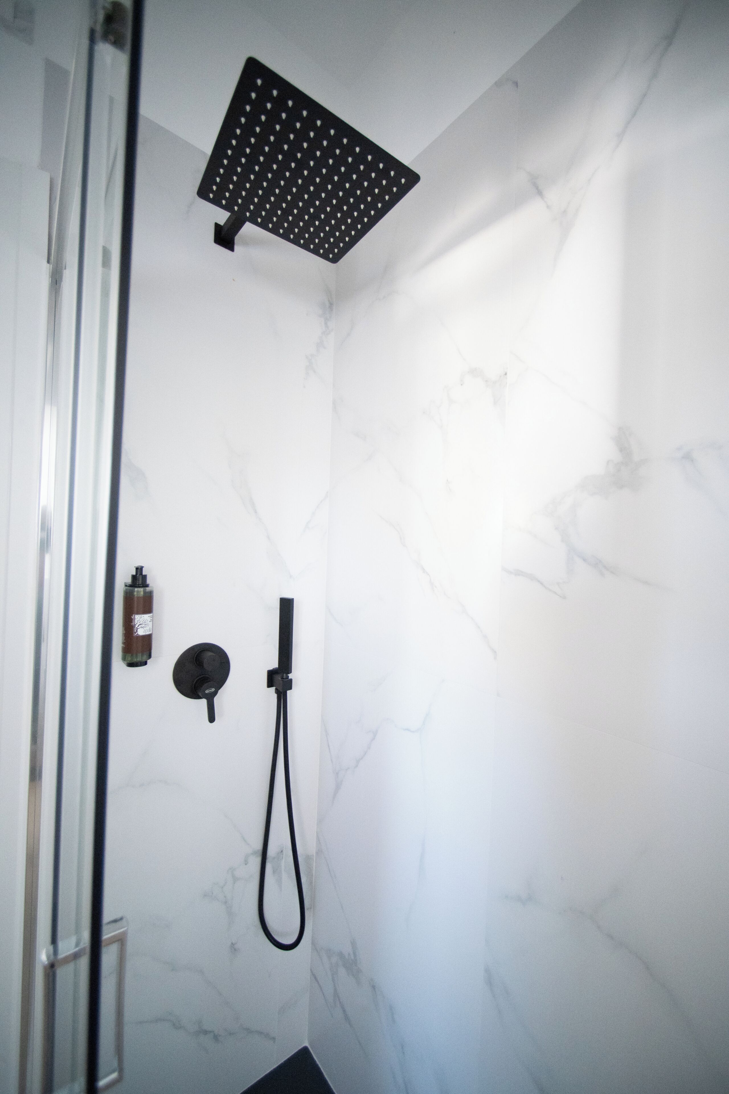 A shower with a black rain showerhead, a handheld showerhead, and a bottle of shampoo on the wall.