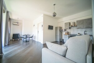 Una sala de estar moderna con sofá, una mesa de comedor con sillas, una zona de cocina con armarios y electrodomésticos, y un televisor montado en la pared.
