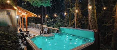 A hot tub in a forested area with a wooden deck and chairs.