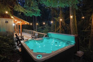 A hot tub in a forested area with a wooden deck and chairs. - The Shuksan | Hot Tub + Game Room + Pool (Deming)