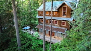 A two-story wooden cabin with a metal roof, surrounded by dense forest. - The Shuksan | Hot Tub + Game Room + Pool (Deming)