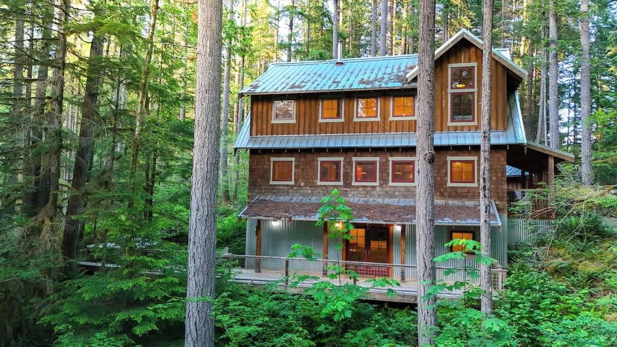 A multi-story cabin in a forest, with a balcony and a visible deck area.