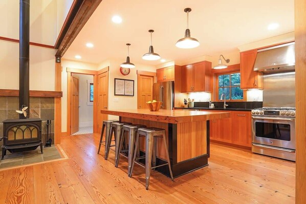Une cuisine avec des meubles en bois, un îlot central doté de tabourets, une cuisinière et des luminaires suspendus.