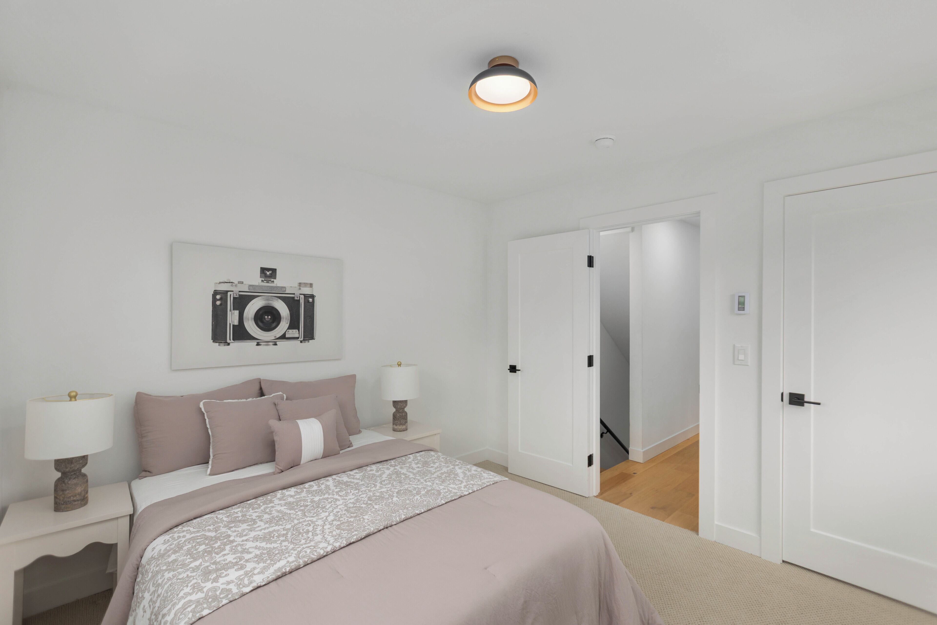 Een slaapkamer met een bed, nachtkastjes, een lamp, een lijst met een afbeelding en een deur naar een andere kamer.