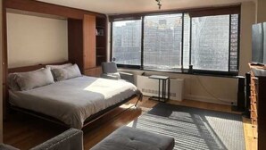 Une chambre à coucher comprenant un lit, un bureau, une chaise, un radiateur et une grande fenêtre avec des stores.