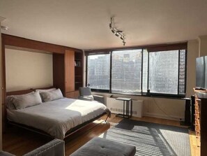 Ein Schlafzimmer mit Bett, Schreibtisch, Stuhl, Heizkörper und einem großen Fenster mit Jalousien.