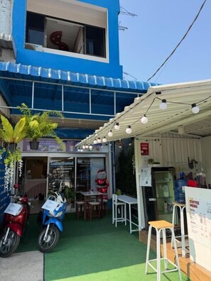 Uma pequena área externa com mesas e cadeiras, um edifício azul, uma motocicleta e um suporte com cardápio de café.