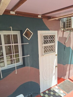 Ein Zimmer mit weißer Tür, einem Fenster mit Metallgitter und einer Wand mit strukturierter Oberfläche.