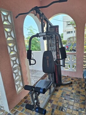 Ein Fitnessraum mit einem Gerät der Marke Marcy, Fliesenboden und Blick auf eine Wohngegend durch ein rundbogenförmiges Fenster.