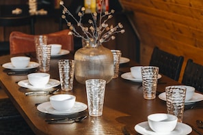 A wooden dining table set with white bowls, plates, and clear glasses, with a vase of branches in the center. - Lakeview A-Frame with hot tub near RMNP (Grand Lake)