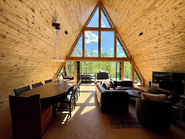 A spacious loft-style room with a high ceiling, a dining area, a seating area with a sofa and a TV, and large windows offering a view of the outdoors.