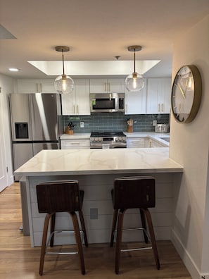 A modern kitchen with white cabinetry, stainless steel appliances, a marble island, and pendant lighting.
