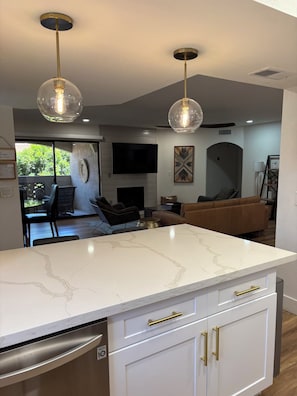 A modern kitchen with a marble countertop, white cabinetry, and gold handles. There is a dining area with a round table and chairs, a sofa, and a flat-screen TV mounted on the wall.