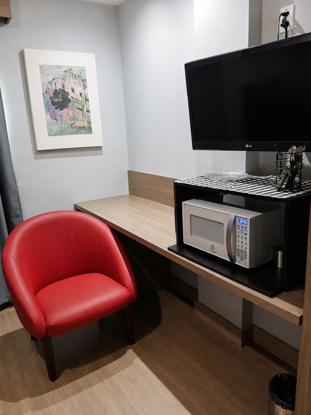 Een kleine kamer met een bureau, een magnetron, een flatscreen televisie, een rode stoel en een ingelijste foto aan de muur.