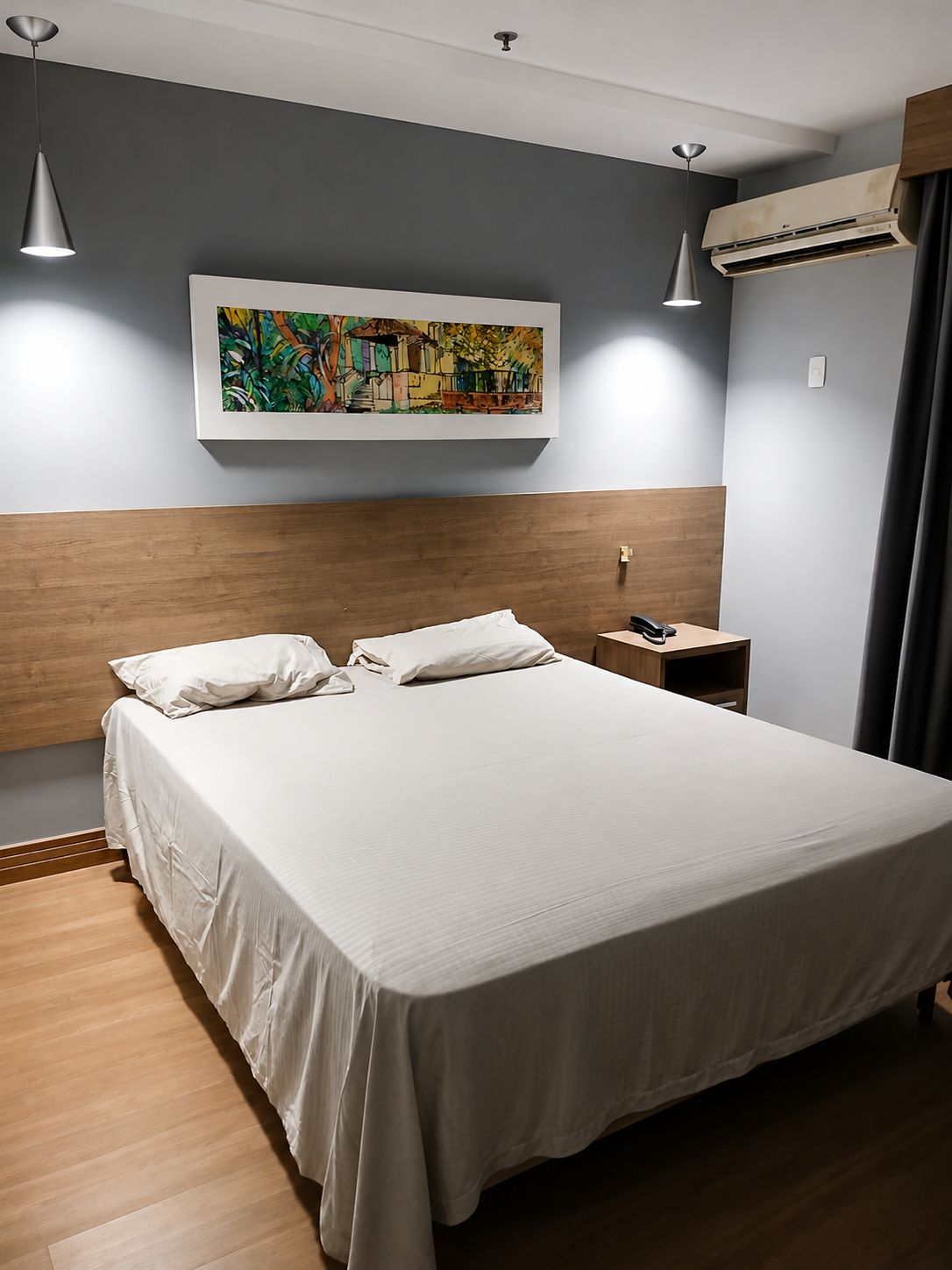 Une chambre d’hôtel moderne avec un grand lit, une tête de lit en bois, une table de chevet, un appareil de climatisation et un tableau au mur.