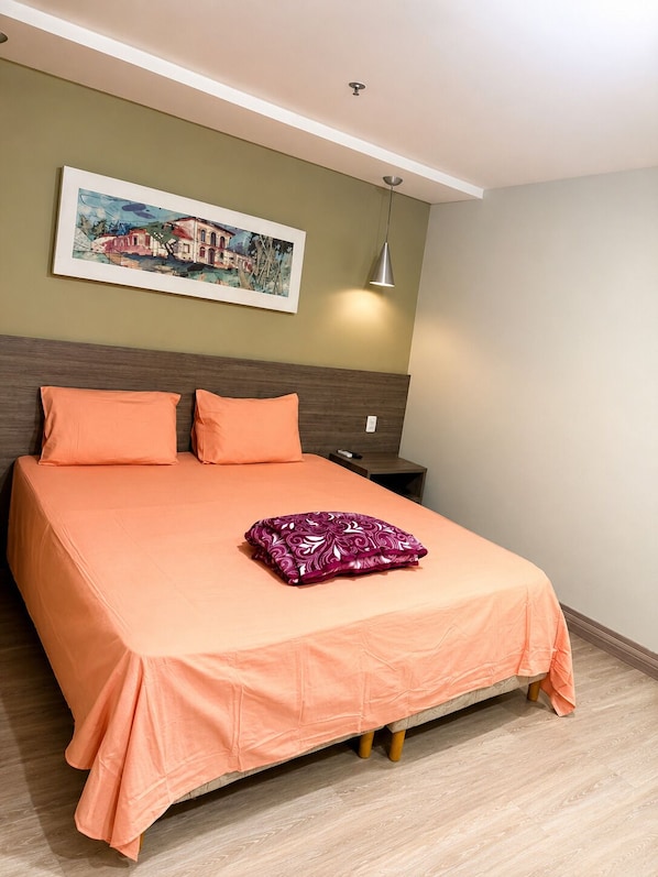 A neatly made bed with a matching orange bedspread, a purple decorative pillow, a wall-mounted painting, and a bedside lamp.