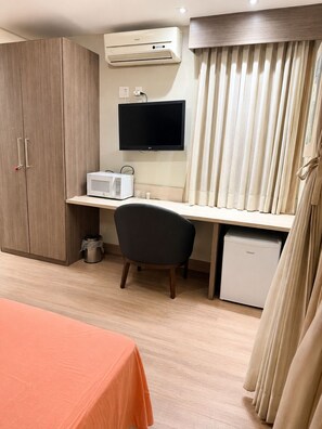 Ein Hotelzimmer mit einem Holzschrank, einem Schreibtisch mit Flachbildfernseher, einer Mikrowelle, einem Sessel, einer kleinen Kühlbox und einem Bett mit orangefarbener Bettdecke.