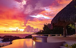 Une vue sur le coucher de soleil depuis un complexe hôtelier doté d’une structure à toit de chaume et d’une piscine en forme de courbe.